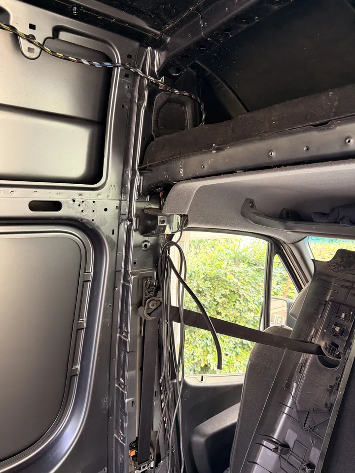 The B-pillar with its trim off. Seatbelt reel exposed and a bundle of cables hanging free.