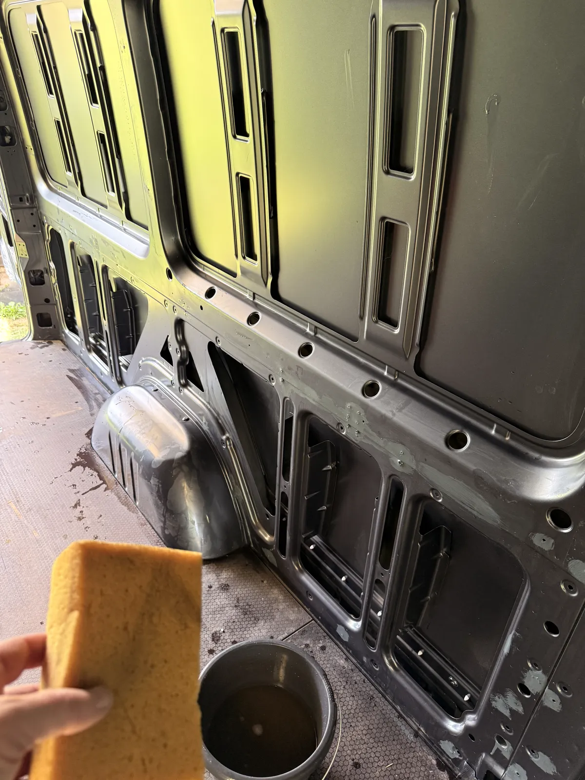 A hand holding a sponge in front of the washed van wall, with a bucket of water on the floor